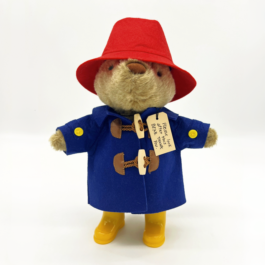 Plush Paddington Bear wearing a red hat, blue duffle coat, and yellow boots, with a tag reading, "Please look after this BEAR. Thank you.