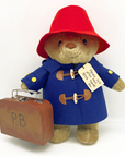 A plush Paddington Bear in a red hat, blue duffle coat, and yellow buttons, holding a suitcase labeled "P.B." with a tag that reads, "Please look after this BEAR. Thank you