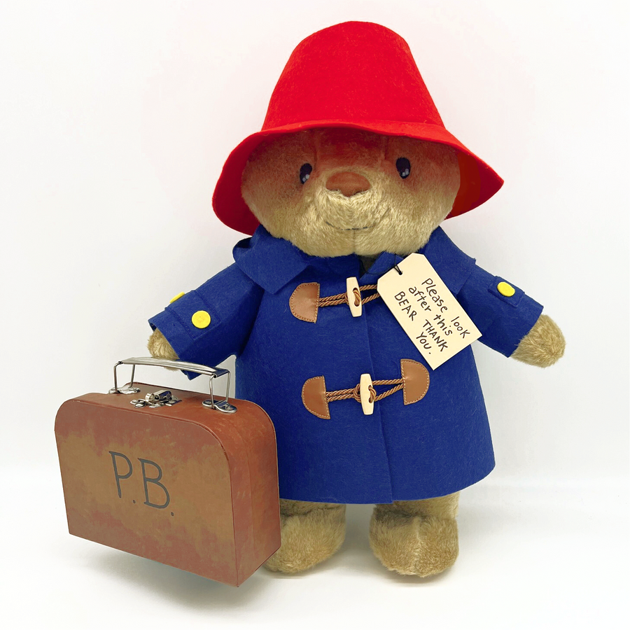 A plush Paddington Bear in a red hat, blue duffle coat, and yellow buttons, holding a suitcase labeled "P.B." with a tag that reads, "Please look after this BEAR. Thank you