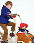 A boy in a blue duffle coat rides a cream bicycle with a wooden cargo box carrying a plush Paddington Bear, a book, and a striped blanket