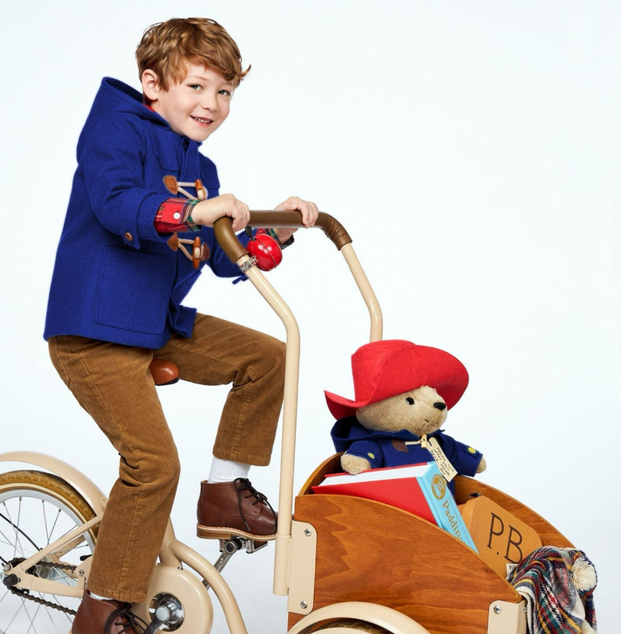 A boy in a blue duffle coat rides a cream bicycle with a wooden cargo box carrying a plush Paddington Bear, a book, and a striped blanket