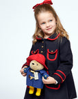 A young girl in a navy coat with red accents holds a plush Paddington Bear, smiling softly 