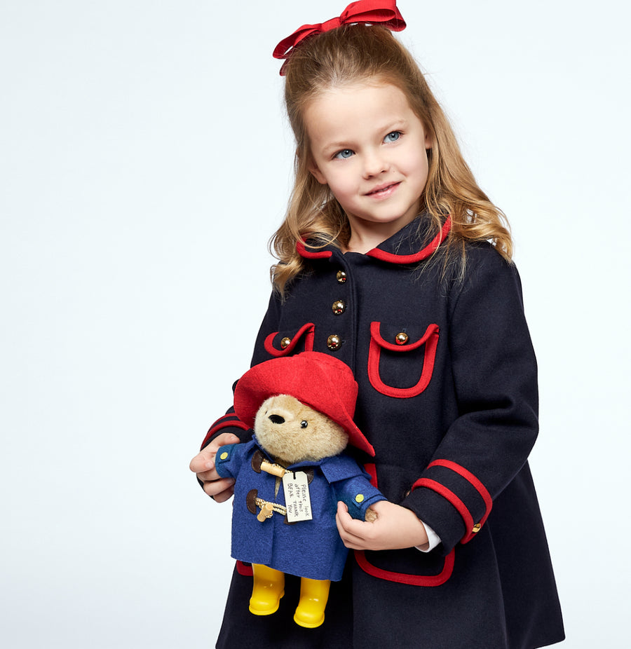 A young girl in a navy coat with red accents holds a plush Paddington Bear, smiling softly 