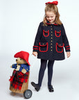 A girl in a navy coat with red accents pulls a wicker pram carrying a plush Paddington Bear wrapped in a red plaid blanket. She smiles while looking down at her toy