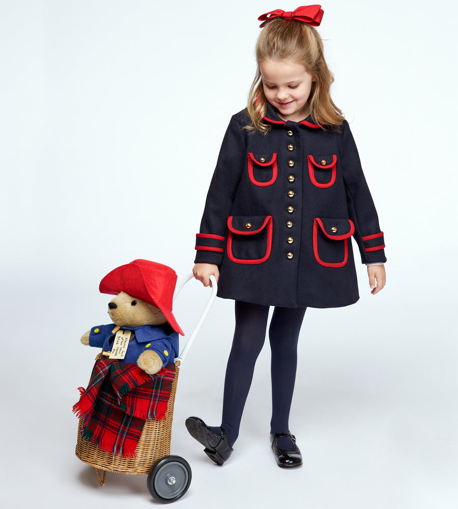 A girl in a navy coat with red accents pulls a wicker pram carrying a plush Paddington Bear wrapped in a red plaid blanket. She smiles while looking down at her toy