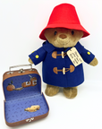 A plush Paddington Bear in a red hat and blue duffle coat stands beside an open suitcase with a blue illustrated interior. A tag on its coat reads, "Please look after this BEAR. Thank you." Set against a white background blue coat