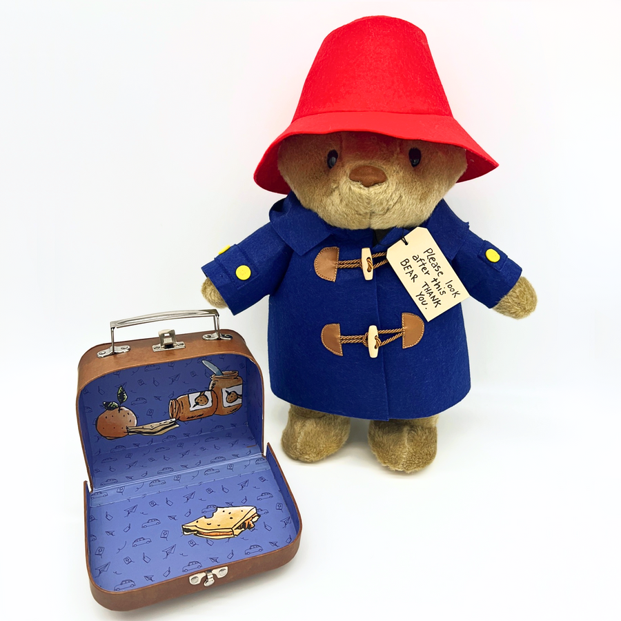 A plush Paddington Bear in a red hat and blue duffle coat stands beside an open suitcase with a blue illustrated interior. A tag on its coat reads, "Please look after this BEAR. Thank you." Set against a white background blue coat