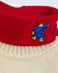 Close-up of a red turtleneck featuring an embroidered Paddington Bear design, worn over a n Ivory  knitted sweater