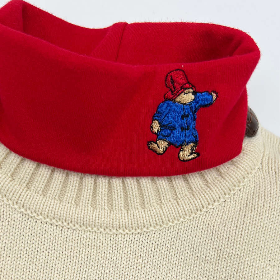 Close-up of a red turtleneck featuring an embroidered Paddington Bear design, worn over a n Ivory  knitted sweater