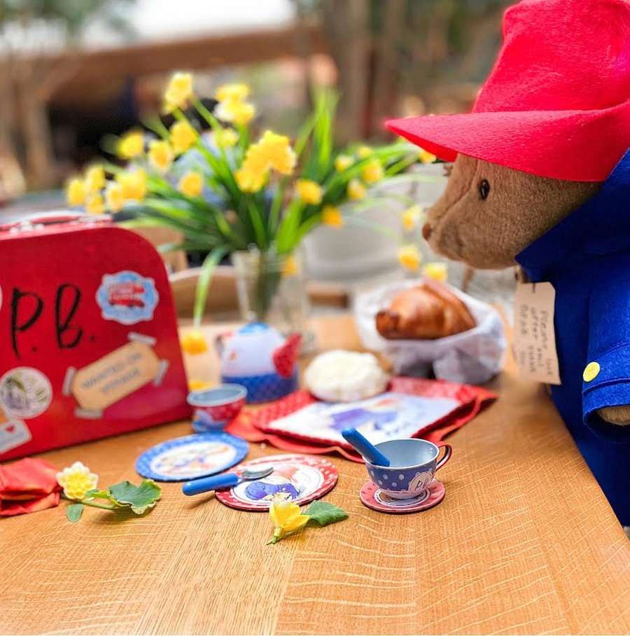 Paddington Bear plush in a red hat and blue coat sits at a tea party table with themed miniature dishes, pastries, and a red suitcase