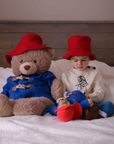 Young boy in a red hat sitting on a bed with two Paddington bear plush toys