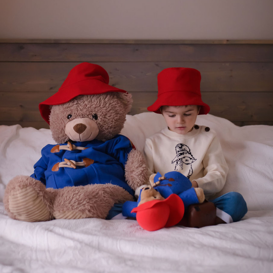 Young boy in a red hat sitting on a bed with two Paddington bear plush toys