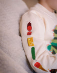 Child wearing a beige-colored sweater with embroidered food illustrations on the left sleeve,