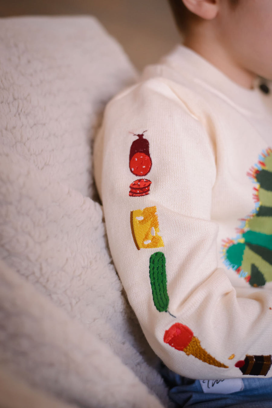 Child wearing a beige-colored sweater with embroidered food illustrations on the left sleeve,