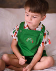 boy wearing a green overall with an embroidered caterpillar design and a short-sleeved button-up shirt featuring a colorful food print.