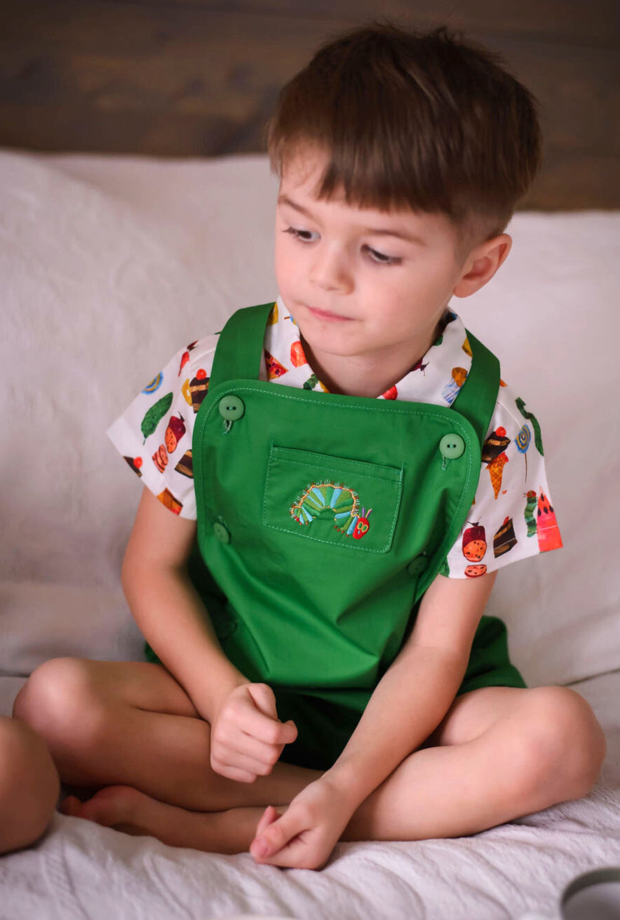 boy wearing a green overall with an embroidered caterpillar design and a short-sleeved button-up shirt featuring a colorful food print.