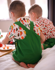 Two young boys sitting on a bed, wearing food-themed print shirts with green overalls, 