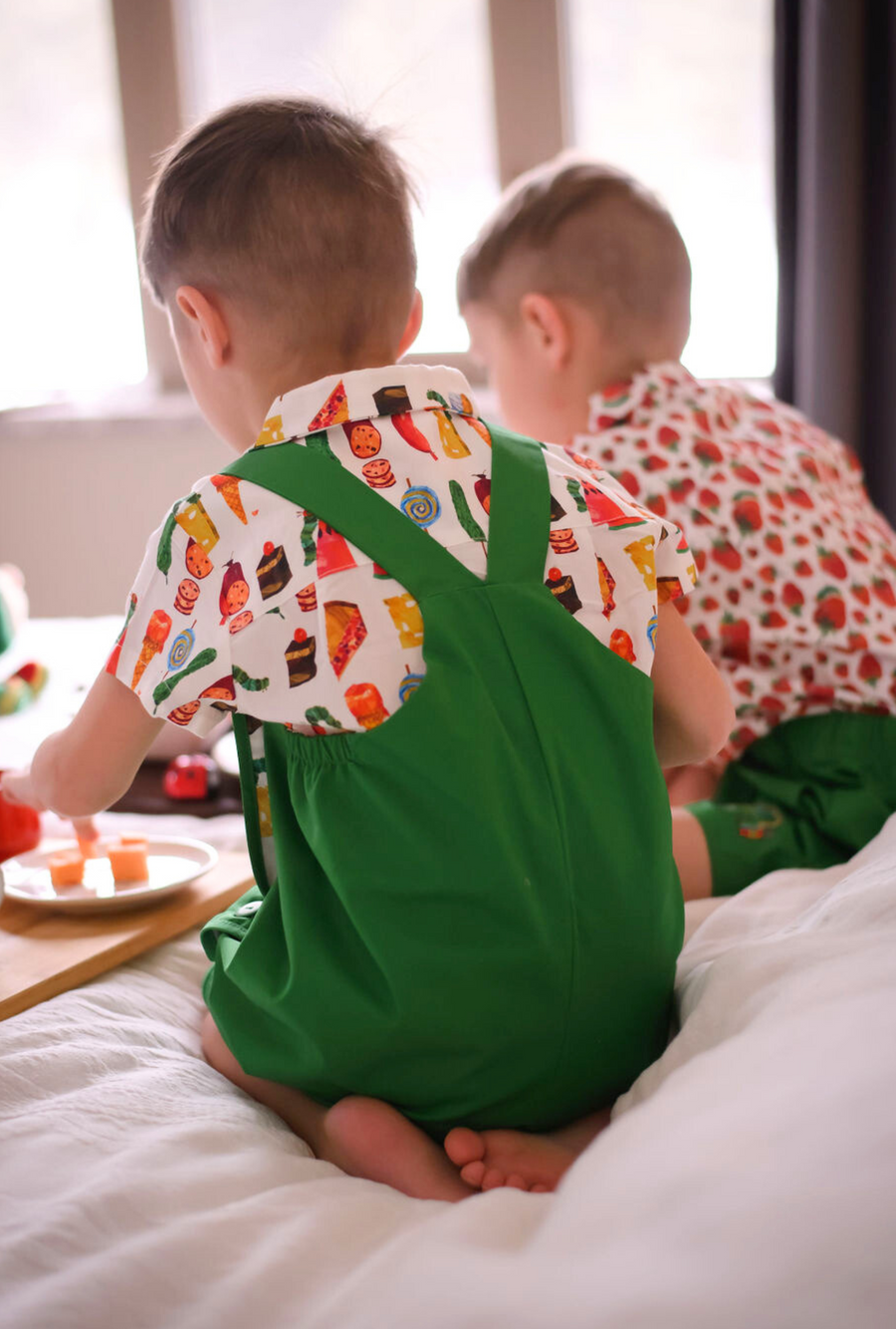 Two young boys sitting on a bed, wearing food-themed print shirts with green overalls, 