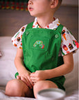 A young child wearing a green romper with an embroidered caterpillar and a food-print shirt, sitting on a bed and looking to the side