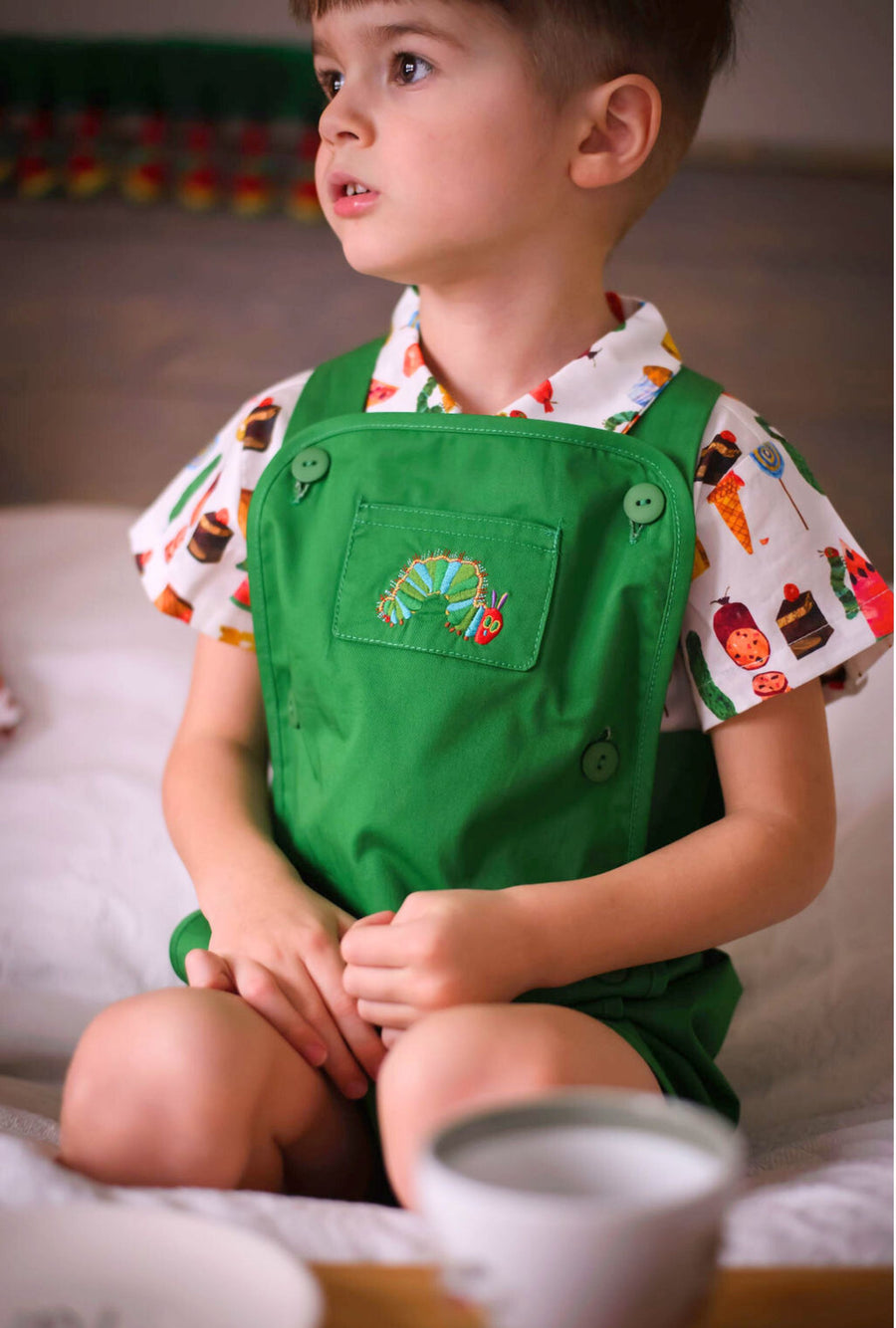 A young child wearing a green romper with an embroidered caterpillar and a food-print shirt, sitting on a bed and looking to the side