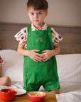 Child in a green romper with a caterpillar embroidery, wearing a food-print shirt in a cozy bedroom.