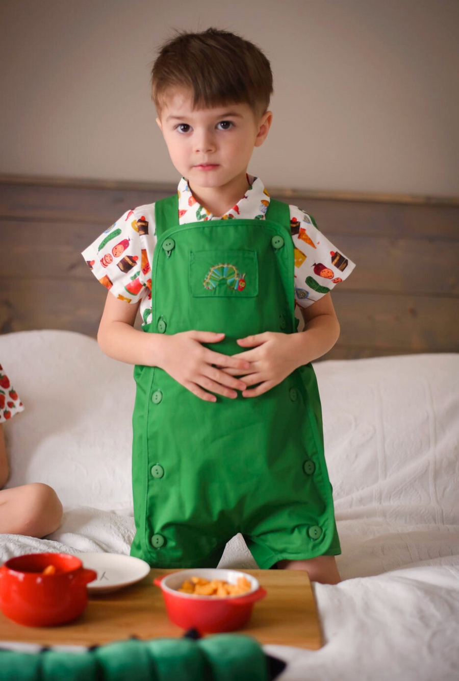 Child in a green romper with a caterpillar embroidery, wearing a food-print shirt in a cozy bedroom.