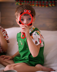 A young child in a green romper and food-print shirt sits on a bed