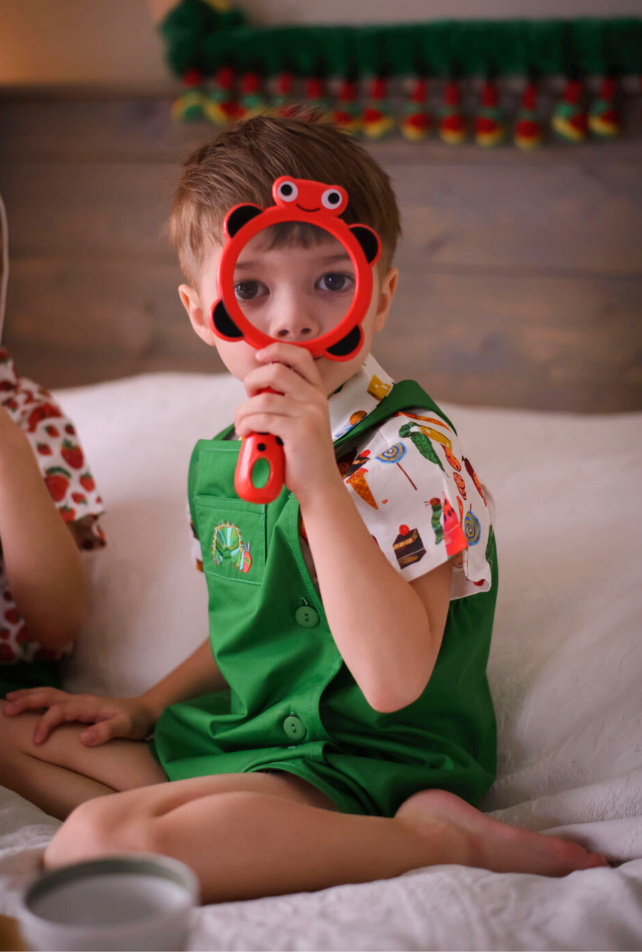 A young child in a green romper and food-print shirt sits on a bed