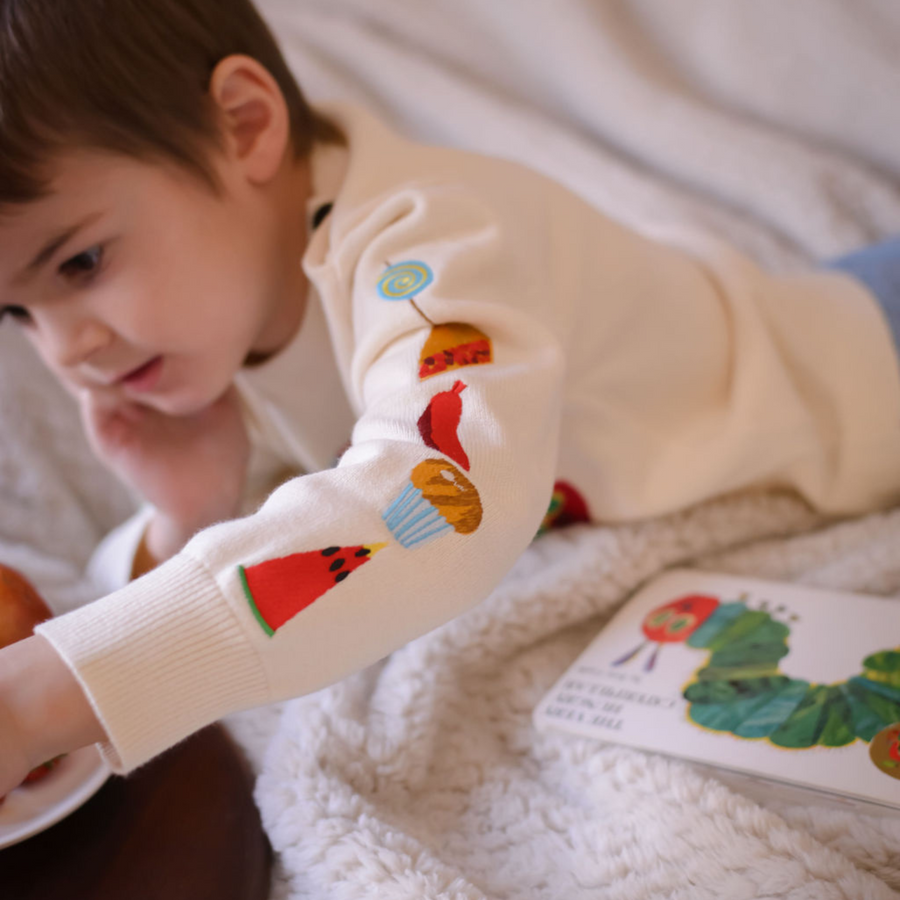 Close-up of a toddler's arm in a Hungry Caterpillar sweater, showing embroidered snacks like watermelon, cupcake, and lollipop