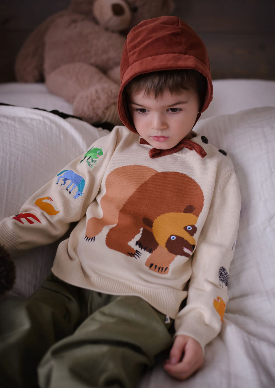 Toddler wearing a soft cotton Brown Bear sweater with embroidered animals on the sleeves, inspired by Eric Carle’s story, sitting on a bed