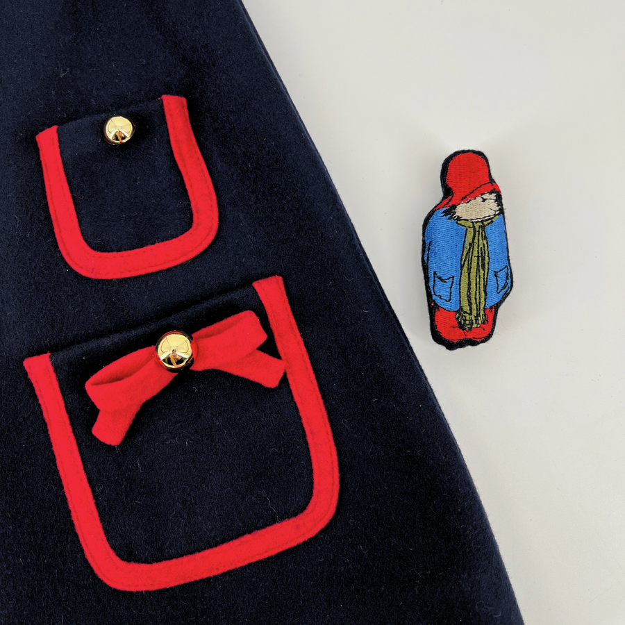 Close-up of a navy blue girls' dress with red-trimmed pockets, gold buttons, and a bow detail, featuring a removable embroidered Paddington Bear