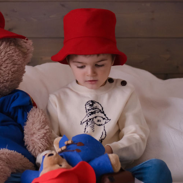 Child wearing a red wool Paddington bucket hat, sitting on a bed with Paddington Bear toys