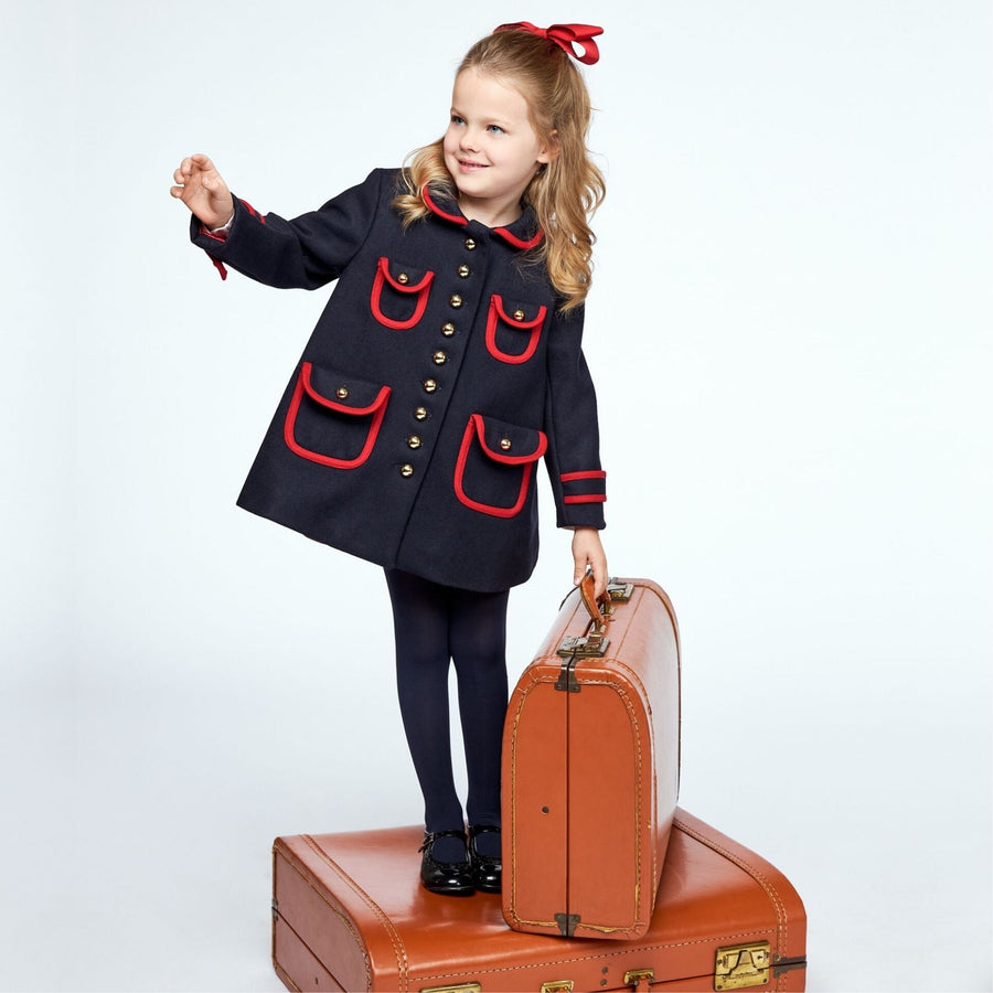 Girl in navy coat with red trim, standing on suitcases