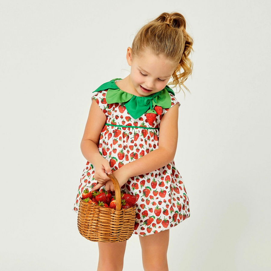 young girl wearing a strawberry-themed dress with a green leaf collar, holding a wicker basket full of fresh strawberries while smiling and looking dow