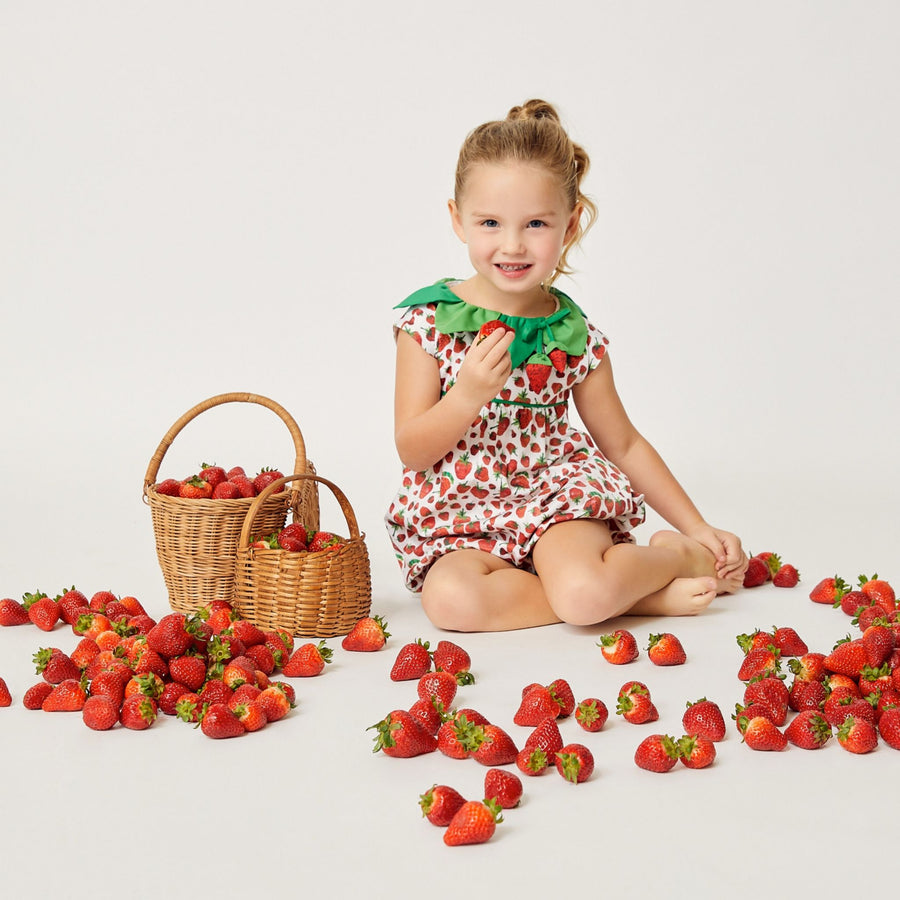  A young girl wearing a strawberry-themed dress sits on the floor surrounded by fresh strawberries