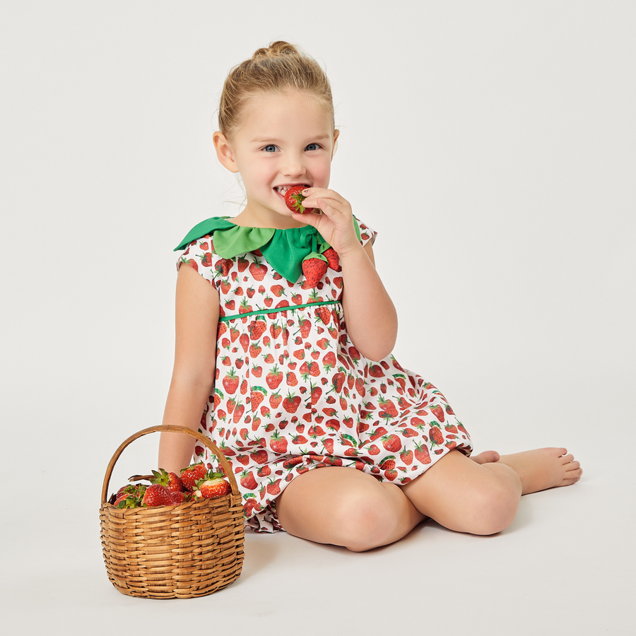 A young girl in a strawberry-patterned dress with a green leaf collar sits on the floor