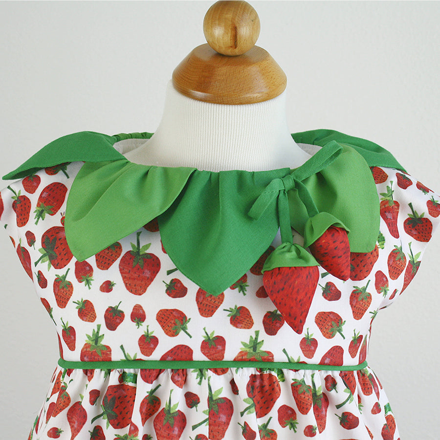 Close-up of a strawberry-themed dress, with red strawberry prints and a green leaf-shaped collar, adorned with two fabric strawberries hanging from a bow