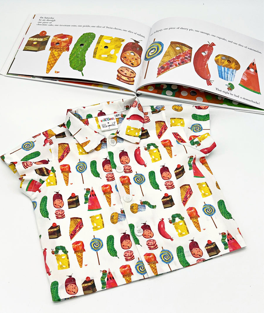 short-sleeved button-up shirt featuring a colorful food-themed print laid flat with an open storybook in the background displaying matching food illustrations