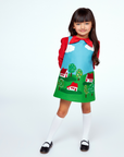 Girl wearing a Little Cloud jumper dress with a red blouse, knee-high socks, and black shoes, featuring appliqué houses, trees, and clouds.