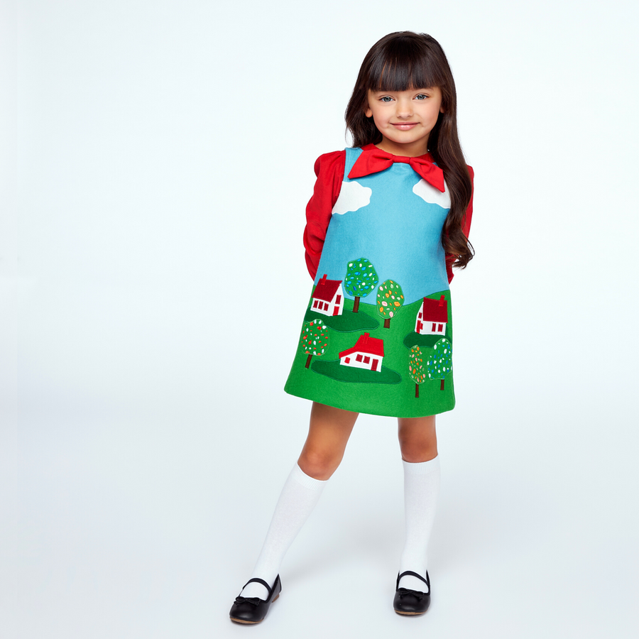 Girl wearing a Little Cloud jumper dress with a red blouse, knee-high socks, and black shoes, featuring appliqué houses, trees, and clouds.